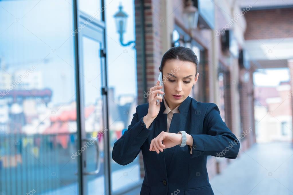 beautiful businesswoman checking time and talking by smartphone on street