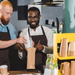 Smiling  young multiethnic owners of coffee shop putting order in paper bag