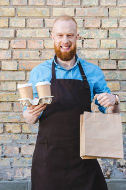 gülümseyen genç erkek barista kağıt bardak kahve karton ve kağıt torbalarda tutan önlük 