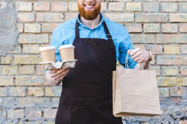 holding kağıt poşet ve tek kullanımlık bardak kahve ile önlük erkek barista gülen in atış kırpılmış