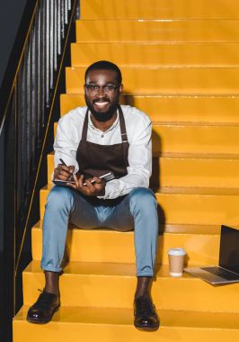 Genç Afro-Amerikan erkek barista önlük holding ders kitabı gülümseyen ve laptop ve kağıt bardak kahve ile merdivenlerde oturan
