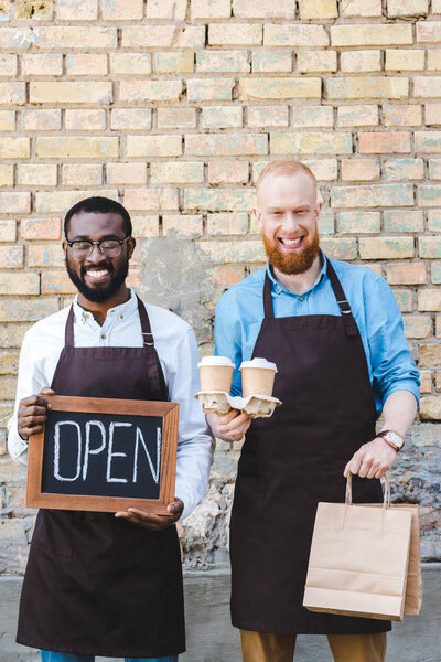 young multiethnic baristas with sign open, paper bags and disposable coffee cups smiling at camera 
