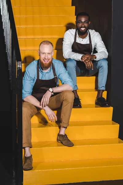 Two Smiling Multiethnic Owners Coffee Shop Aprons Sitting Stairs — Free Stock Photo