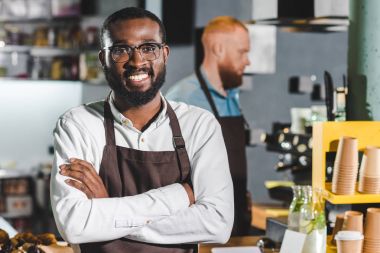 Afro-Amerikan erkek barista önlük arkasında, meslektaşı ile gülümseyen genç portresi 