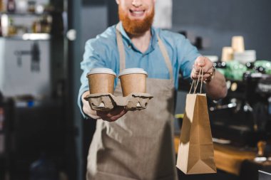 Tek kullanımlık kahve fincanı ve kafede kağıt torba tutarak gülümseyen sakallı barista kırpılmış atış