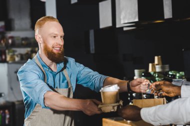 gülen in kırpılmış atış genç barista kafede Afro-Amerikan alıcı kağıt torba ve tek kullanımlık kahve fincanı veren sakallı