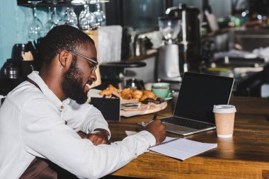 Not alma ve kafede boş ekranlı dizüstü bilgisayar kullanarak gözlük içinde gülümseyen genç Afro-Amerikan barista