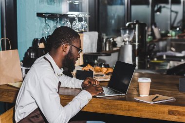 gülümseyen genç Afro-Amerikan barista kahve dükkanında dizüstü kullanarak gözlük içinde