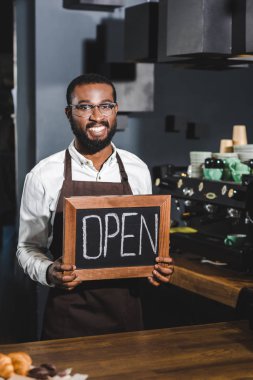 yakışıklı genç Afro-Amerikan barista işareti açık ve gülümseyerek kafede kamera tutan gözlük içinde