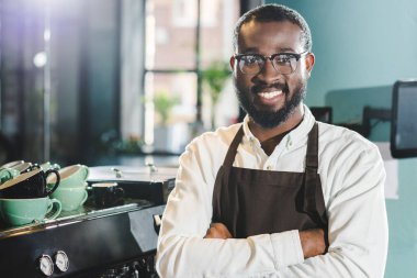 neşeli Afro-Amerikan barista önlük ve çapraz kolları ile ayakta ve kamera Cafe gülümseyen gözlük
