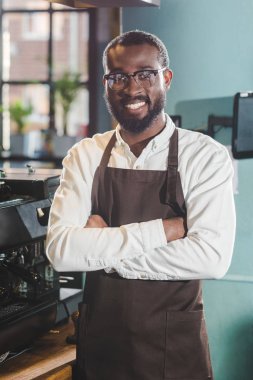 mutlu genç Afro-Amerikan barista ayakta çapraz kolları olan ve kamera kafede gülümseyen