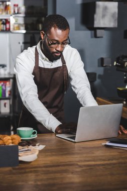 Genç Afro-Amerikan barista kafede çalışırken dizüstü bilgisayar kullanarak