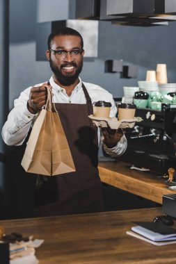 handsome smiling young african american barista holding disposable coffee cups and paper bags in coffee shop  clipart