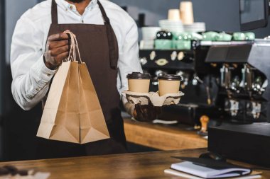 cropped shot of african american barista holding disposable coffee cups and paper bags in cafe