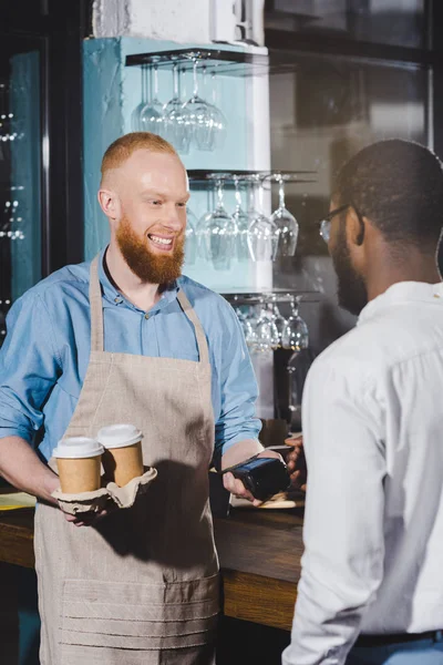 kredi kartıyla ödeme ve terminal ve kağıt bardak kahve ile erkek barista gülümseyen bir Afrikalı-Amerikalı adam