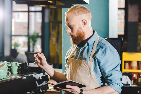 young male barista standing in coffee shop with textbook 