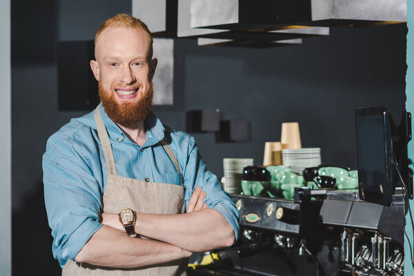 confident bearded barista in apron standing with crossed arms and smiling at camera in coffee shop