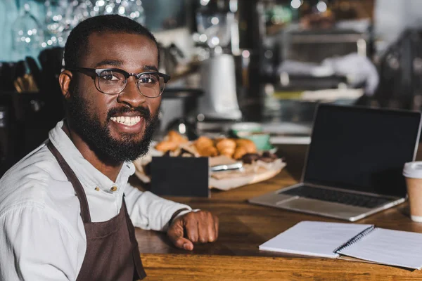 カフェで働きながらカメラで笑顔の眼鏡でハンサムな若いアフリカ系アメリカ人のバリスタ — ストック写真