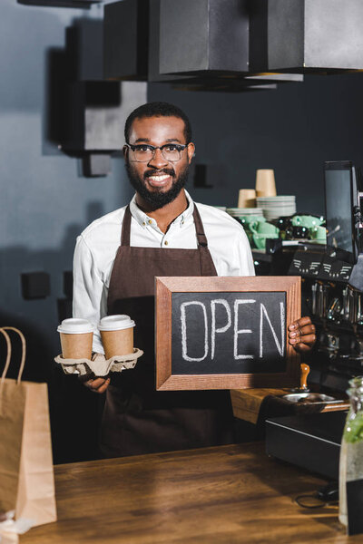 young african american barista in eyeglasses holding sign open and disposable paper cups, smiling at camera in coffee shop