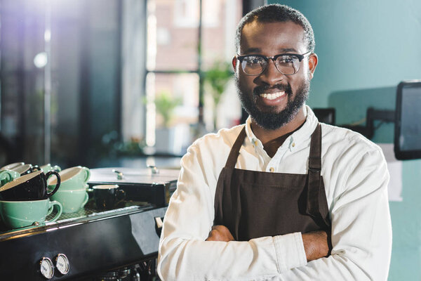 cheerful african american barista in apron and eyeglasses standing with crossed arms and smiling at camera in cafe