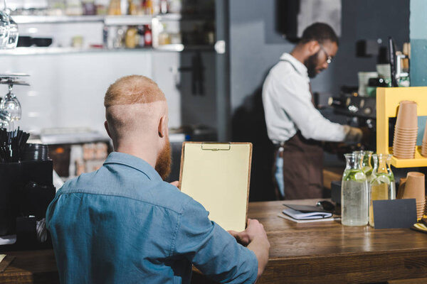 rear view of young man holding blank clipboard while barista making coffee behind 
