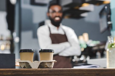 İki kağıt bardak kahve ve Afro-Amerikan barista ayakta arkasında çapraz silah ile gülümseyen yakından görmek