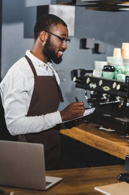 smiling young african american barista in eyeglasses taking notes while woeking in cafe clipart