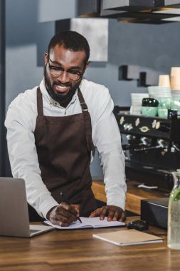 Not alma ve kahve dükkanında dizüstü kullanarak gülümseyen genç Afro-Amerikan barista 
