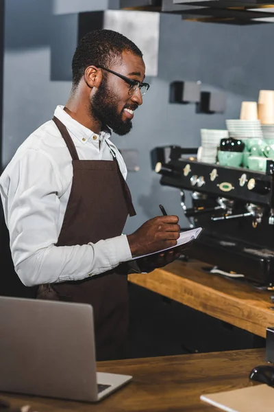 Uśmiechnięty Młody Afroamerykanin Barista Okularach Sporządzania Notatek Podczas Woeking Kawiarni — Zdjęcie stockowe