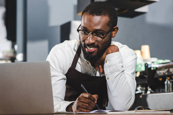 handsome young african american barista taking notes and using laptop at coffee shop  