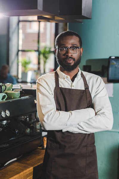 handsome african american barista standing with crossed arms and looking at camera in coffee shop 