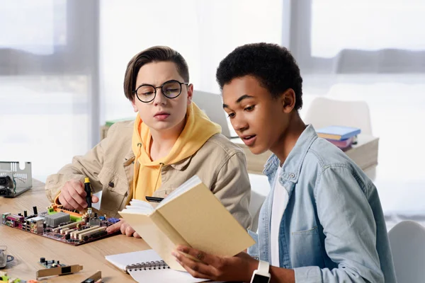 Multicultural Teen Boys Reading Book Soldering Motherboard Home — Stock Photo, Image