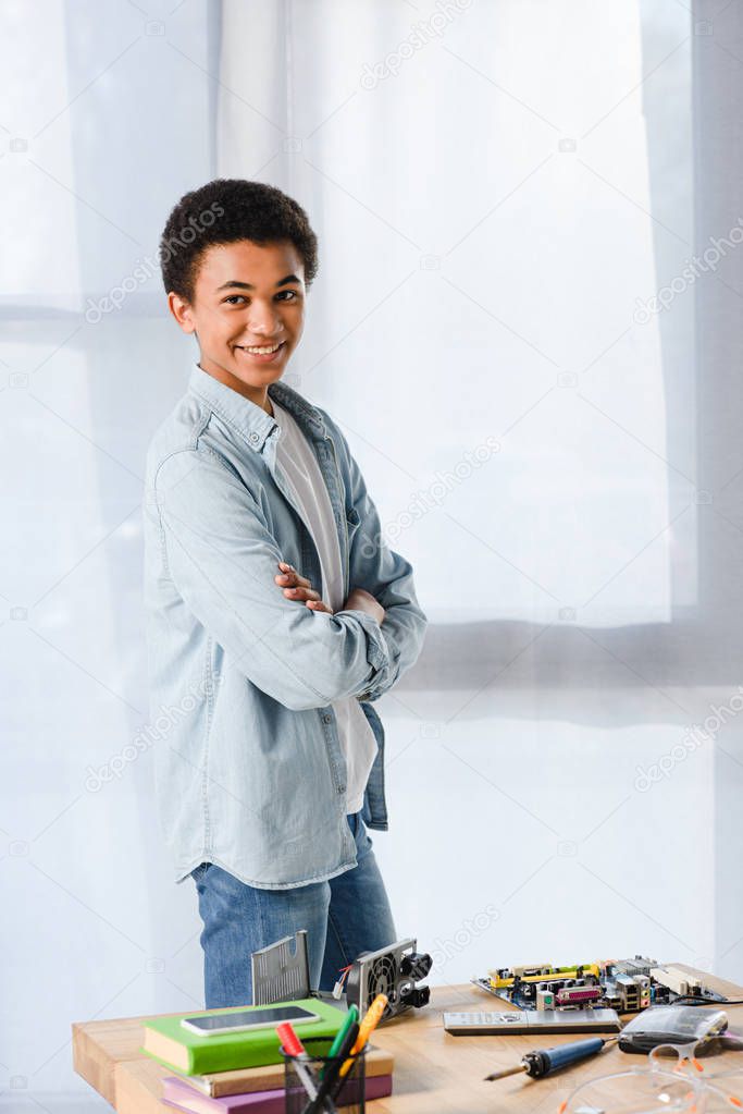 smiling african american teenager standing with crossed arms near table with equipment at home