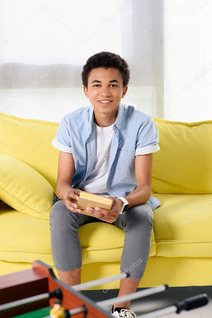 smiling african american teenager sitting with book on yellow sofa at home