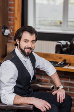 handsome barber sitting on chair at workplace and looking at camera clipart