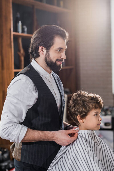 handsome barber looking at reflection of little kid sitting in chair at barbershop
