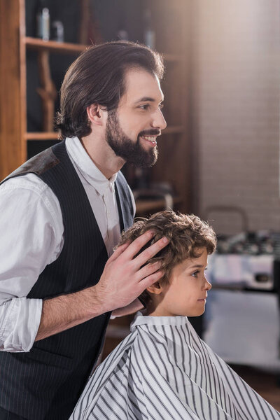 smiling handsome barber looking at reflection of little kid sitting in chair at barbershop