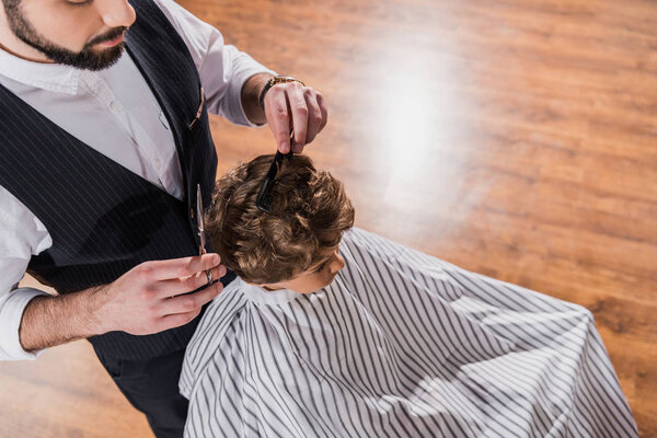 high angle view of kid covered with striped cloth sitting at barbershop while barber cutting his hair