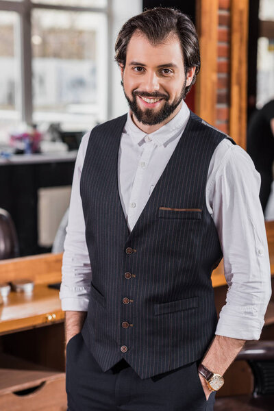 handsome young barber in stylish vest looking at camera in barbershop