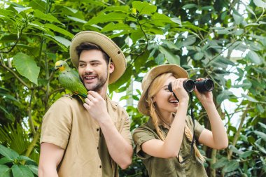 happy young couple with parrot sitting on shoulder of man hiking together in jungle clipart