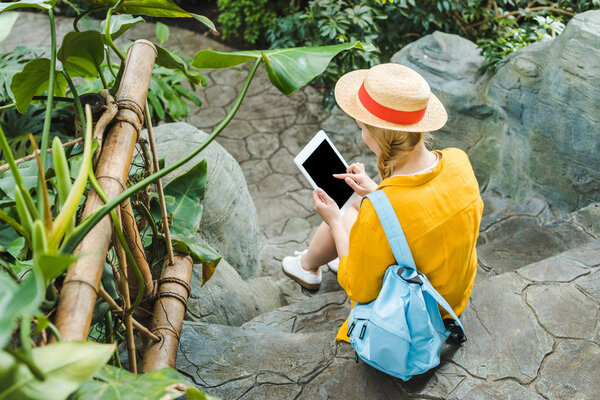 rear view of young woman in straw hat using tablet while sitting on stone staircase in park