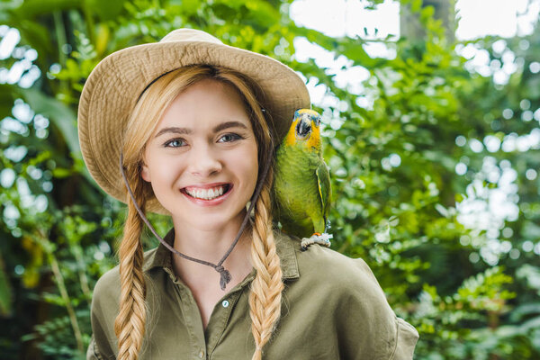 happy young woman in safari suit with parrot on shoulder in jungle