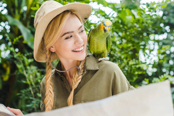 smiling young woman in safari suit looking at parrot on shoulder while navigating in jungle with map