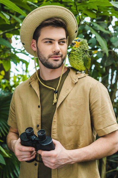 handsome young man in safari suit looking at parrot on shoulder in jungle