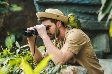 handsome young man with parrot on shoulder looking through binoculars in jungle