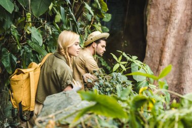 active young couple in safari suits hiking in jungle and looking somewhere impressed clipart