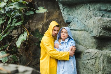 frozen young couple in raincoats embracing and trying to warm up under rocks in jungle clipart
