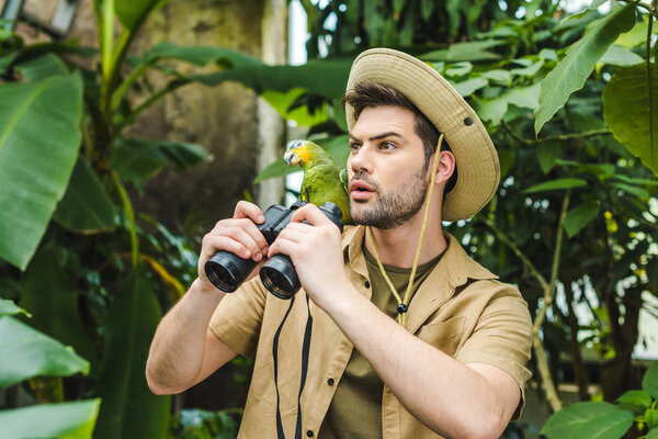 shocked young man with parrot on shoulder and binoculars in jungle