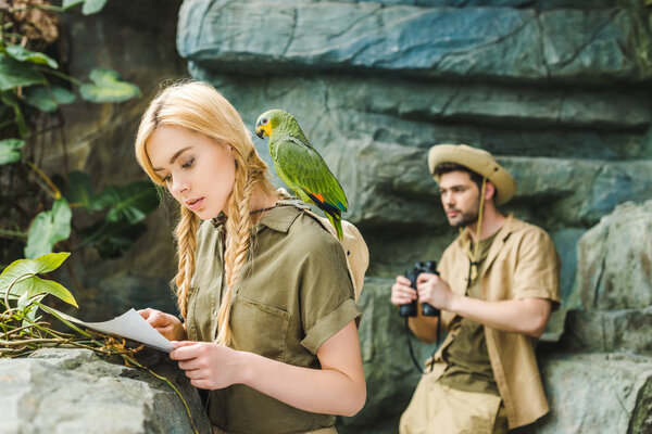 attractive young woman in safari suit with parrot and map navigating in jungle while her boyfriend looking through binoculars