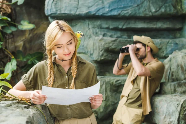 young woman in safari suit with parrot and map navigating in jungle while her boyfriend looking through binoculars